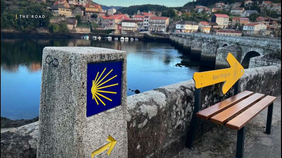 A stone Camino marker with a scallop shell and a yellow arrow beside a harbour, chapter card reading The Road
