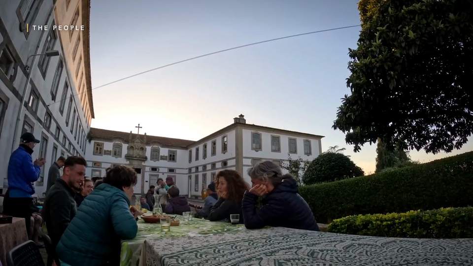 Pilgrims sharing a meal at a long outdoor table in front of a monastery, chapter card reading The People