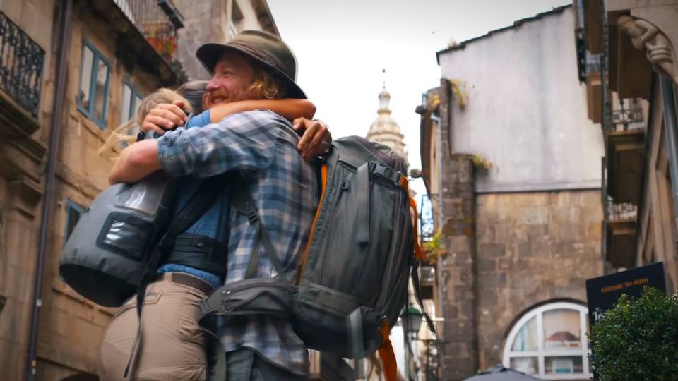 Two pilgrims with backpacks hugging in a Santiago street, the cathedral tower behind them