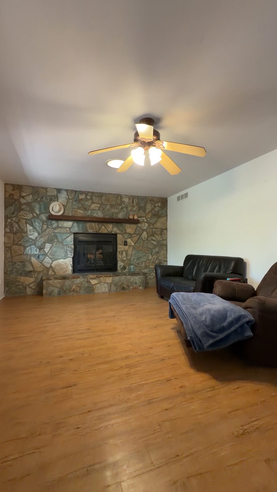 The living room as filmed: stone fireplace, ceiling fan, plank floor, with the seller's leather recliners and a storage bin in the room